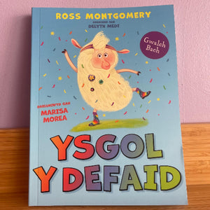 Book titled 'Ysgol y Defaid' with colorful cover and character illustration on a pink background
