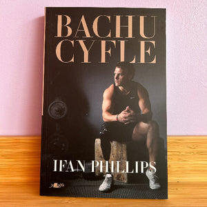 Book titled 'Bachu Cyfle' by Ifan Phillips on a wooden surface with a pink wall background.