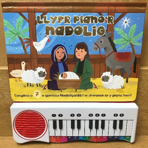 A children's activity book titled 'Llyfr Piano'r Nadolig' with a colorful cover featuring a cartoon illustration of a donkey, a sheep, and two people singing. There is a small electronic piano keyboard below the image.