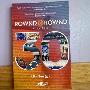 Book titled 'Rownd Rownd yn dathlu'r' with a colorful cover on a wooden surface.