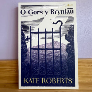 Book cover of 'O Gors y Bryniau' by Kate Roberts on a wooden surface with a purple wall background.