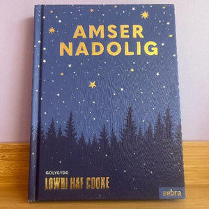 A book titled 'Amser Nadolig' with a dark blue cover featuring stars, trees, and the name 'Lowri Haf Cooke' as the editor.
