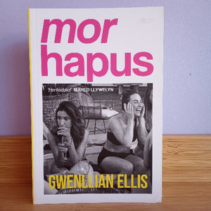 Book titled 'mor hapus' by Gwenllian Ellis on a wooden surface with a purple wall background.