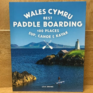 Book cover of 'Wales Cymru Best Paddle Boarding 100 Places' featuring an image of a paddle boarder on a body of water with a lighthouse and mountain in the background.