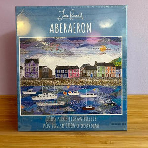 A puzzle box with a scenic puzzle piece design featuring a seaside town, labelled as a 1000 piece jigsaw puzzle by Josie Russell.