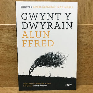 Paperback book cover of 'Gwynt y Dwyrain' by Alun Ffred featuring a black and white image of a tree against a stormy sky.