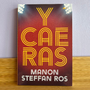 Book cover of 'Y CAE RAS' by Manon Steffan Ros on a wooden surface with a light purple wall background.