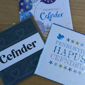 Three greeting cards with 'Cefnder' and 'PENBLWYD HAPUS CEFNDER' text on a wooden surface.
