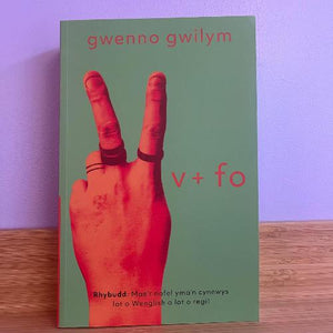 A book cover featuring a hand making a peace sign with a green background, with the title 'V + Fo' and the author's name 'GWENNO GWILYM' at the top.