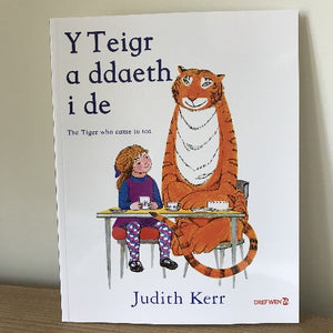 Cover of the book 'Y Teigr a ddaeth i de - The Tiger who came to tea' by Judith Kerr, depicting an illustration of a young girl, a tiger, and a table.