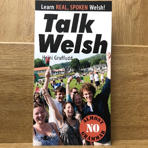 The cover of the book 'Talk Welsh' by Heini Gruffydd features a group of people smiling and waving at an outdoor event, with the title and author's name displayed prominently.