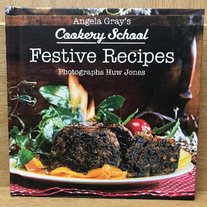A book cover of 'Angela Gray's Cookery School: Festive Recipes' featuring a photograph of a roasted meal against a backdrop with a lit flame and red checkered pattern.