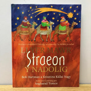 A book cover of 'Llyfr Mawr Straeon y Nadolig' featuring illustrations of people on camels against a desert backdrop, with a starry night sky above.
