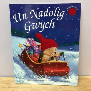 A book cover of 'Un Nadolig Gwych' featuring an illustration of a hedgehog in a red sled with Christmas presents, set against a snowy background.