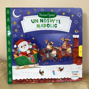 A colorful book cover of 'Un Noswyl Nadolig' featuring illustrations of Santa Claus, reindeer, and Christmas decorations against a starry night background.