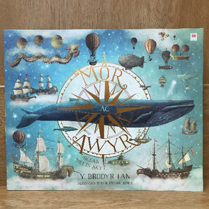 Cover of the book 'Môr ac Awyr - y Brodyr Fan' featuring illustrations of a whale, ships, balloons, and a compass rose against a blue background with a pattern of stars and a whale.