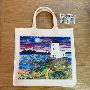 Tote bag with a scenic design of a lighthouse and landscape on a wooden surface.
