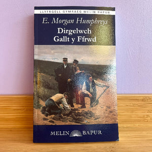 Book cover of 'Dirgelwch Gallt y Ffrwd' by E. Morgan Humphreys on a wooden surface with a purple wall background.