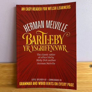 Book cover of 'Bartleby Yr Ysgriffennwr' by Herman Melville on a wooden surface with a white wall background.