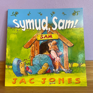 The children's book 'Symud, Sam!' by Jac Jones, featuring a colorful illustration of a person and a dog under a wooden house, resting on a wooden surface against a purple wall.