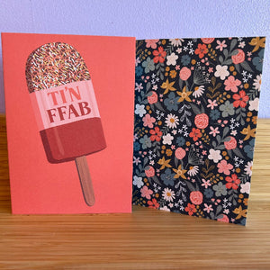 Two greeting cards on a wooden surface with one featuring a ice cream cone and 'TIN FFAB' text, the other with floral pattern.