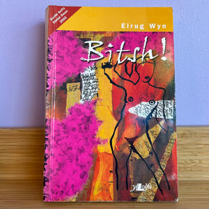 Book titled 'Bitsh!' by Eirug Wyn on a wooden surface with a purple wall background