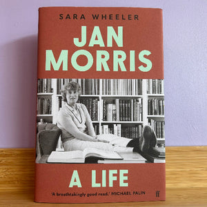 Book cover of 'Jan Morris: A Life' by Sara Wheeler on a wooden surface with a purple wall background.