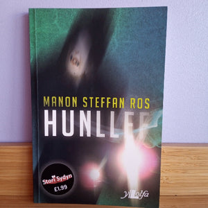 Book titled 'Hunllef' by Manon Steffan Ros on a wooden surface with a light purple wall background.