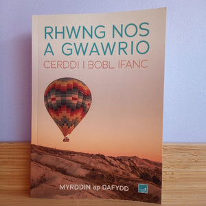 Book titled 'RHWNG NOS A GWAWRIO' with a hot air balloon on the cover, placed on a wooden surface with a light blue wall background.
