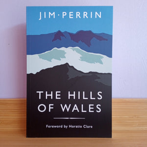Book cover of 'The Hills of Wales' by Jim Perrin on a wooden surface with a light purple background