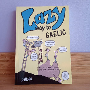 Book titled 'Lazy Way to Gaelic' with cartoon illustrations on a wooden surface and purple wall background.