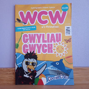 Welsh magazine cover titled 'WCW a'i ffrindiau' featuring colorful cartoon illustrations including a penguin, a blue shark, a smiling sun, and a butterfly, photographed standing on a wooden surface against a light purple wall.