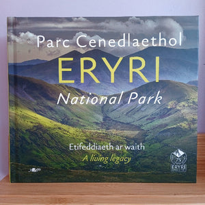 A hardcover book titled 'Eryri National Park' resting on a wooden surface against a light purple wall. The cover features a scenic landscape of green and blue mountains under a cloudy sky, with the title 'ERYRI' printed in large yellow letters.