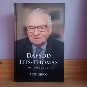 The book 'Dafydd Elis-Thomas: Nation Builder' by Aled Eirug, standing upright on a wooden surface against a light purple wall. The cover features a portrait of the subject wearing glasses and a suit.