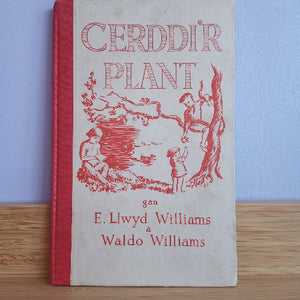 A vintage hardcover book titled 'CERDDIR PLANT' by E. Llwdyd Williams and Waldo Williams, featuring red text and illustrations on an off-white cover, standing upright on a wooden surface against a light blue wall.