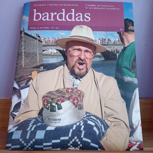 A physical copy of the Welsh magazine 'barddas' (Haf 2026) is shown lying on a wooden surface against a light purple wall. The cover features a man in a beige suit and a cream-colored fedora hat, holding a patterned textile with a white hat resting on top.