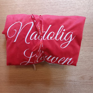 A folded red textile item, likely a blanket or throw, with white cursive text reading 'Nadolig Llwen' tied with a red ribbon, placed on a light wood surface.