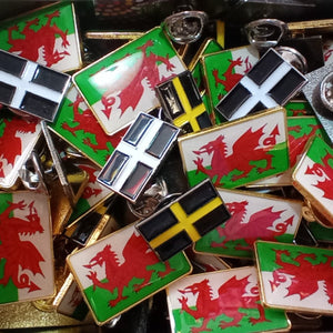 A pile of small rectangular enamel pins featuring Welsh flag designs, including the red dragon on a green and white background, as well as black and white cross flags and a black and yellow flag.