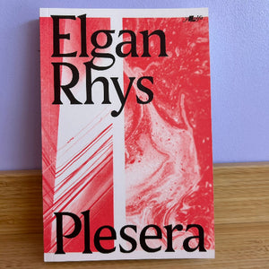 Book cover of 'Plesera' by Elgan Rhys with red and white design on a wooden surface and blue wall background.
