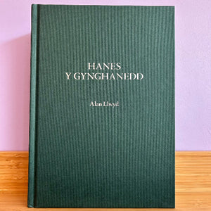 Green book titled 'Hanes Y Gynghganedd' by Alan Llwyd on a wooden surface with a pink background