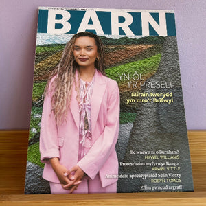 A copy of the BARN magazine, featuring a woman in a pink blazer on the cover, displayed standing on a wooden surface against a light purple wall.