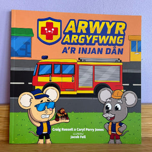 A children's book titled 'ARWYR ARGYFWG A'R INJAN DÃN' featuring cartoon mouse characters and a fire truck illustration on the cover, photographed standing on a wooden surface against a light purple wall.