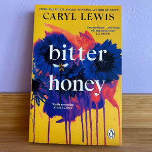 The book cover of 'bitter honey' by Caryl Lewis, featuring a bright yellow background with blue and orange paint-dripping floral artwork and a bee, photographed standing on a wooden surface against a light wall.