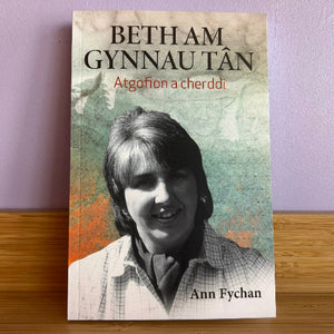 A book titled 'Beth Am Gynau Tân' by Ann Fychan, featuring a portrait of Ann Fychan on the cover, displayed on a wooden surface against a light purple wall.