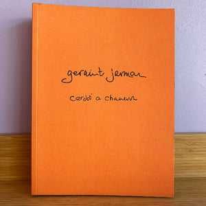 An orange book standing upright on a wooden surface against a light gray wall. The cover has black handwritten text reading 'geraint jarman' and 'Cerddi a chaneuon'.