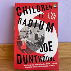 The book cover of 'Children of Radium' by Joe Dunthorne, featuring a bold red and white design with black text and a central black-and-white photograph of children holding hands. The book is photographed standing upright on a wooden surface against a light purple wall.