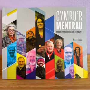 A book titled 'CYMRU'R MENTRAU' by Menna Jones, featuring a colorful collage of portraits on the cover, resting on a wooden surface against a light purple wall.