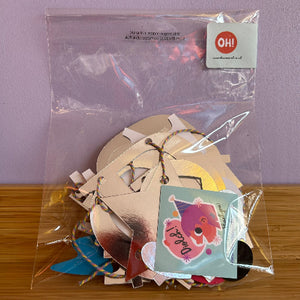A clear plastic bag filled with assorted craft cutouts and small decorative pieces, including metallic gold shapes, a blue piece, and a light blue card with a cartoon face. A white sticker with the brand name 'Oh! Designs' is visible in the top right corner.