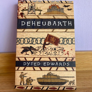A book titled 'Deheubarth' by Dyfed Edwards, featuring a detailed, textured cover illustration in a military campaign style with a tank, soldiers, skulls, and a flag, displayed on a wooden surface against a light purple background.