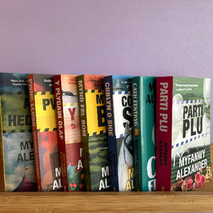 A row of colorful hardcover books standing upright on a wooden shelf against a light purple wall. Visible titles include 'My Alien', 'Party Plu', 'My Fanwy Alexander', and others with authors' names such as Myfanwy Alexander and Cath Fenting.